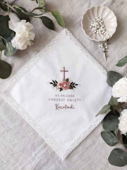 Haftowana szatka z krzyżem i kwiatami oraz imieniem dziecka oraz datą, zdobiona koronką.