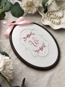 Owalny haftowany obrazek z inicjałem i kwiatami