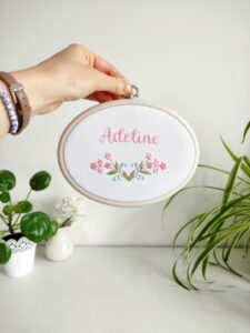 owalny biały obrazek z różowymi i niebieskimi kwiatkami i haftowanym imieniem