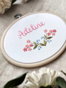 owalny biały obrazek z różowymi i niebieskimi kwiatkami i haftowanym imieniem