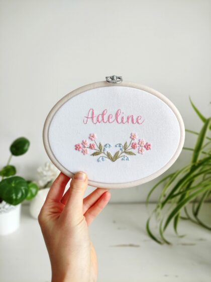 owalny biały obrazek z różowymi i niebieskimi kwiatkami i haftowanym imieniem