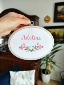 owalny biały obrazek z różowymi i niebieskimi kwiatkami i haftowanym imieniem