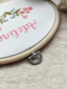 owalny biały obrazek z różowymi i niebieskimi kwiatkami i haftowanym imieniem