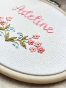 owalny biały obrazek z różowymi i niebieskimi kwiatkami i haftowanym imieniem